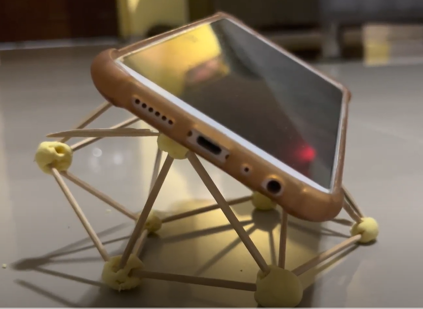 A smartphone in a brown case propped up on a DIY stand made from toothpicks and small yellow clay balls.